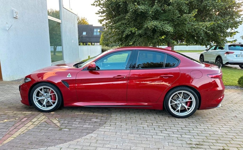 Seitenansicht eines roten Alfa Romeo Giulia Quadrifoglio von der Fahrerseite aus betrachtet. Das Auto steht vor einem Haus mit grüner Wiese. Die Felgen sind silber. Der Wagen ist sehr sauber und glänzt.